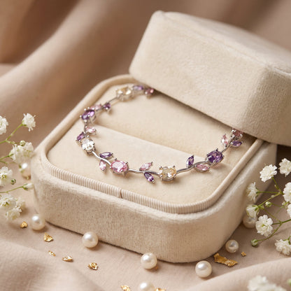 A delicate bracelet with purple, pink, and clear gemstones set in silver-toned metal, displayed on a cream velvet jewelry box surrounded by white baby's breath flowers and scattered pearls.