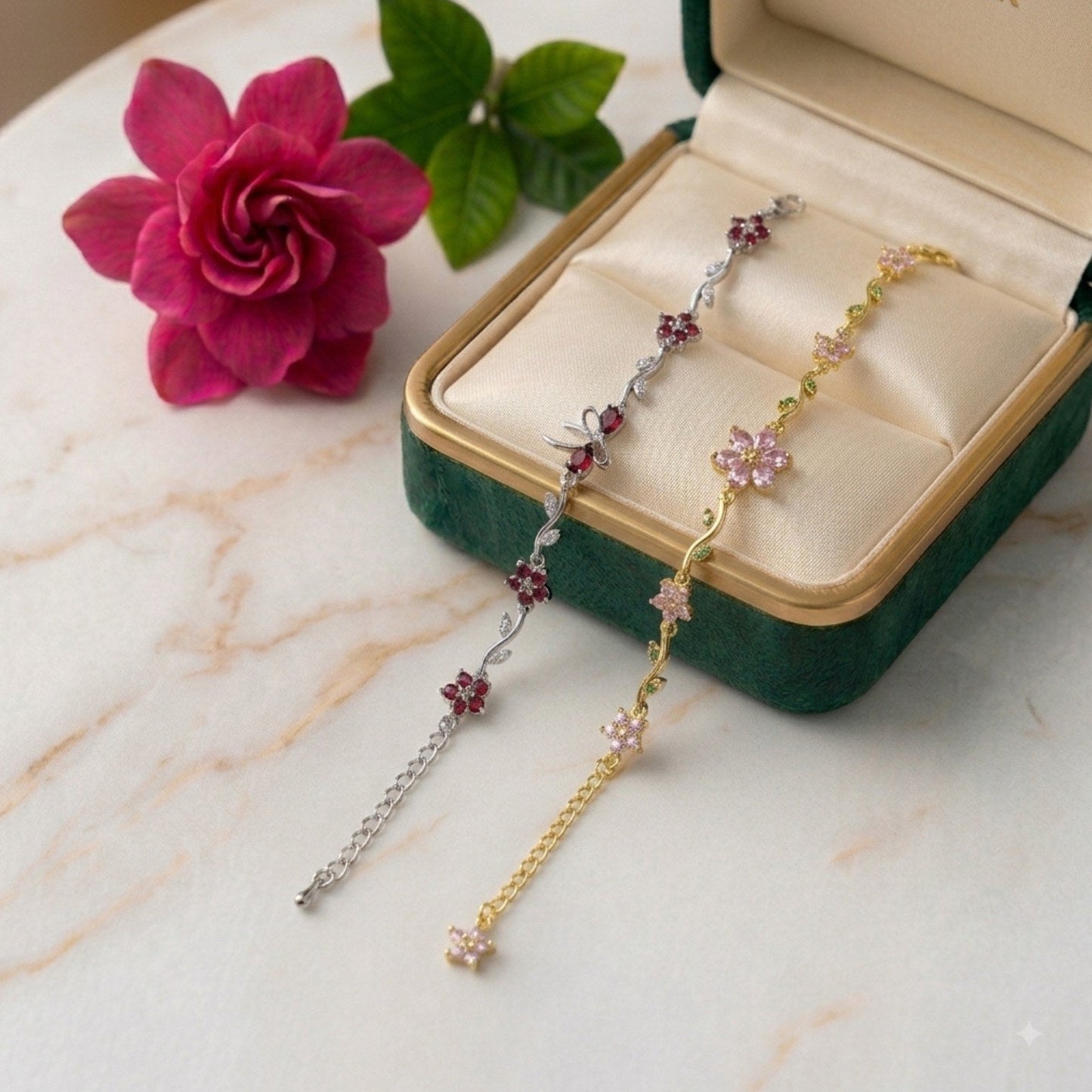 Two delicate floral chain bracelets displayed in an open green jewelry box on a light marble surface, with a pink rose and green leaves beside the box.