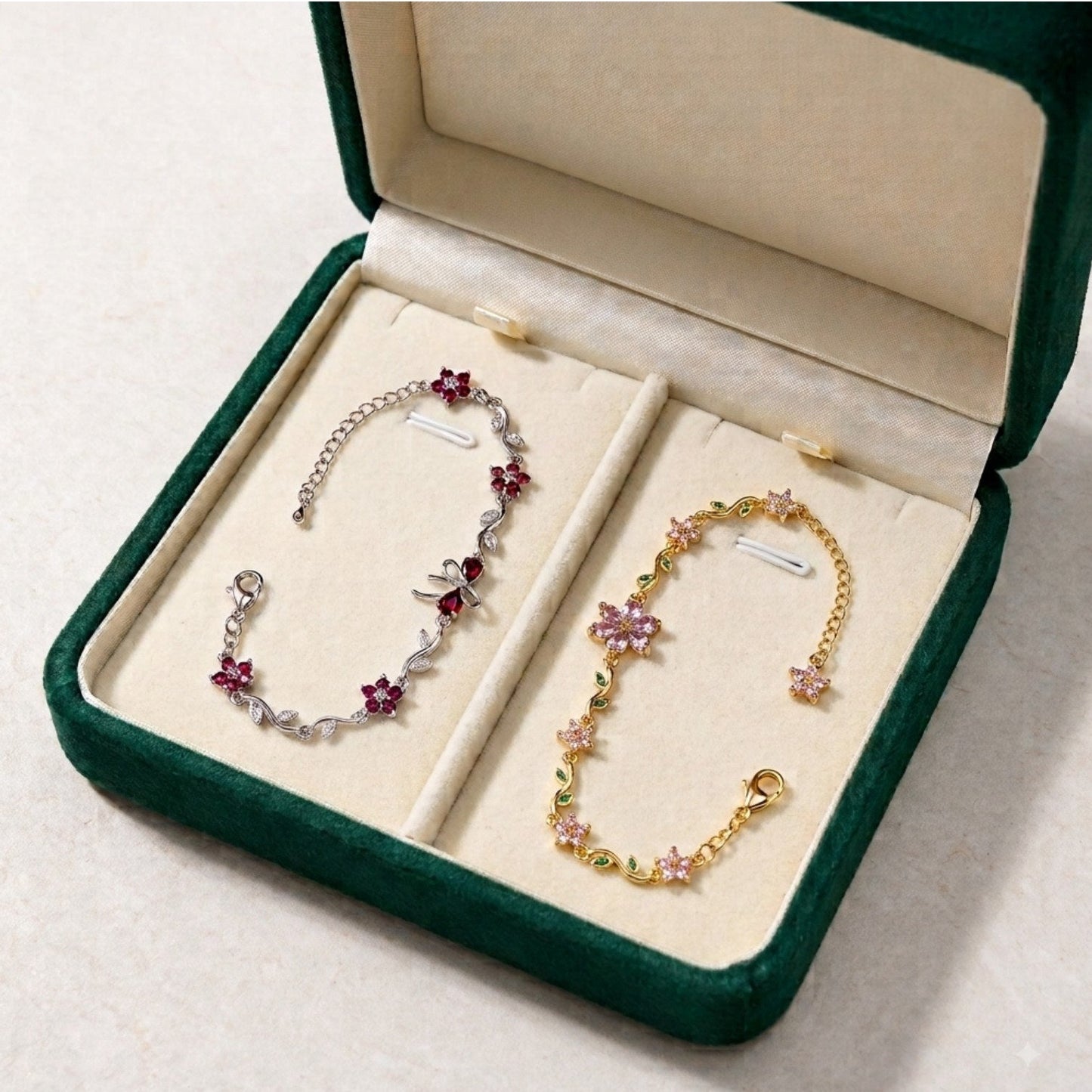 Two delicate floral chain bracelets displayed in an open dark green jewelry box with cream lining. One bracelet is silver-toned with pink flower charms, and the other is gold-toned with pink and green flower charms.