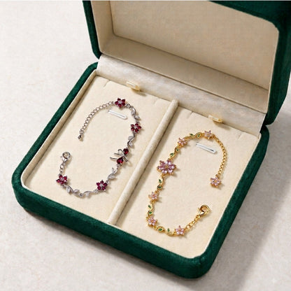 Two delicate floral chain bracelets displayed in an open dark green jewelry box with cream lining. One bracelet is silver-toned with pink flower charms, and the other is gold-toned with pink and green flower charms.
