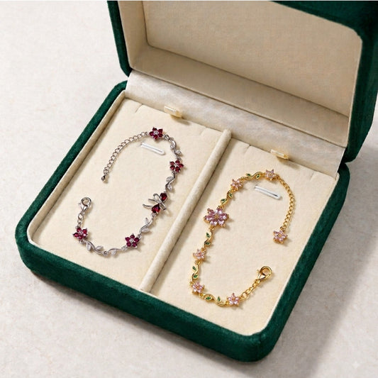Two delicate floral chain bracelets displayed in an open dark green jewelry box with cream lining. One bracelet is silver-toned with pink flower charms, and the other is gold-toned with pink and green flower charms.